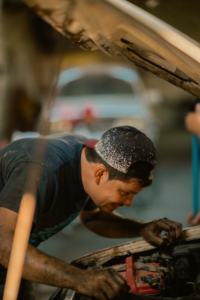 gallery-04 Mechanic repairing car under open hood, focused, wearing cap, selective focus.